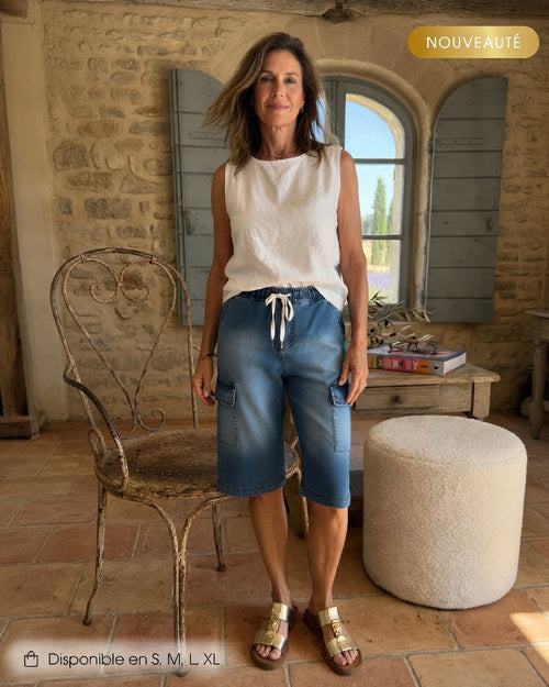 Bermuda cargo en denim bleu moyen à taille élastiquée et cordon blanc, poches cargo latérales, revers bas de jambe, porté avec top blanc sans manches et sandales dorées, mas provençal