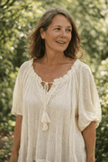 Tunique oversize en filet ajouré écru à col V lacet et pompons, manches bouffantes mi-longues, coupe ample grande taille, portée en jardin verdoyant, lumière dorée estivale