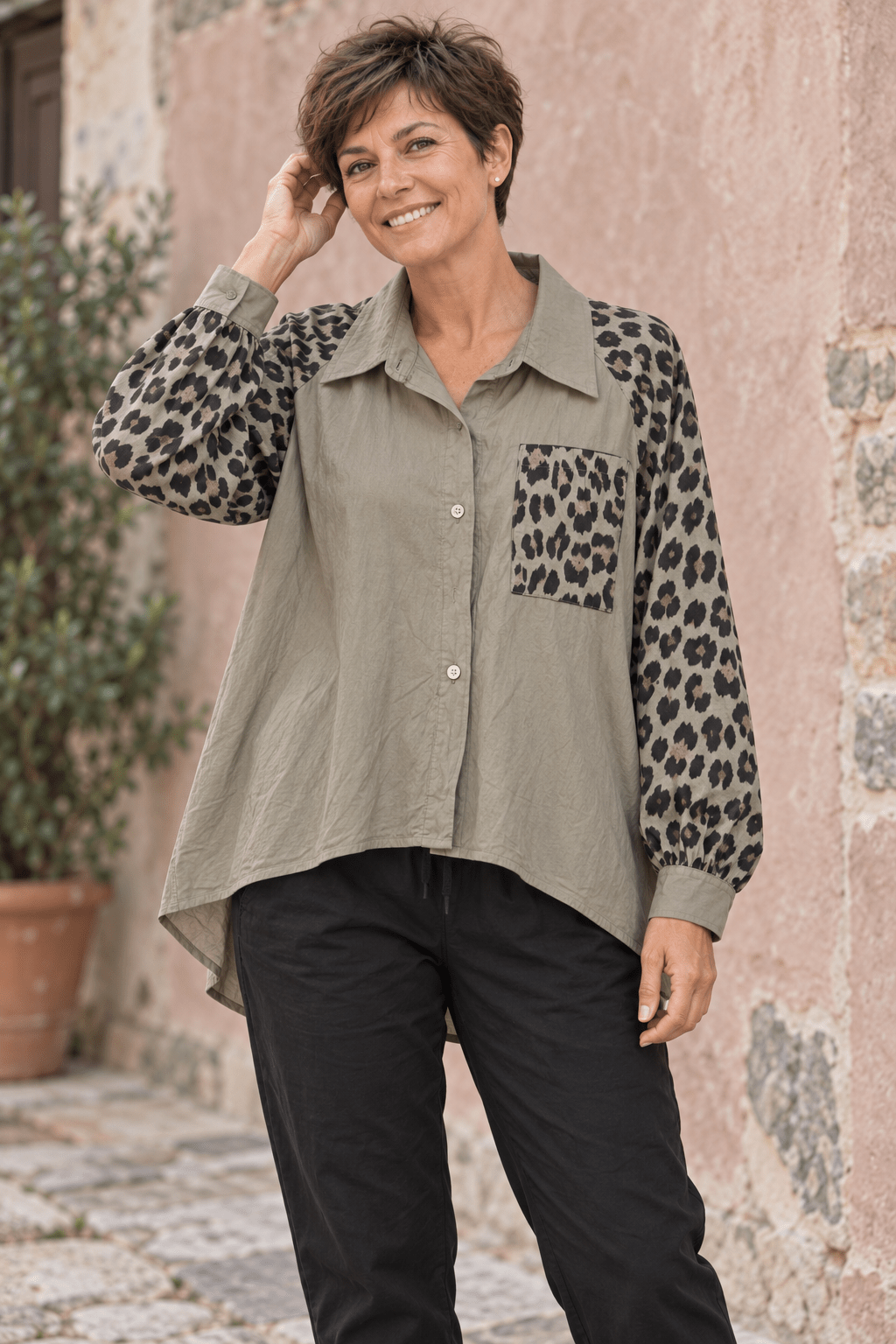 Femme aux cheveux courts portant la chemise taupe à manches et poche léopard avec un pantalon noir, devant un mur vieux rose.