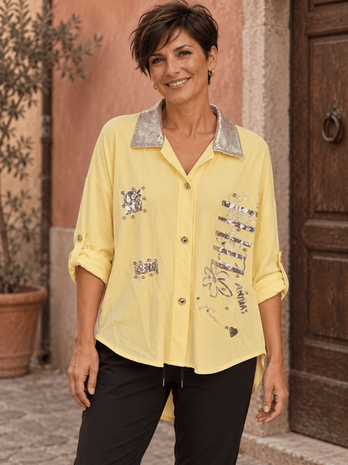Chemise jaune à sequins et strass portée avec un pantalon noir, manches relevées et col métallisé.