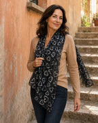 Femme portant le foulard Batticuore noir à cœurs blancs, drapé de façon asymétrique sur l’épaule