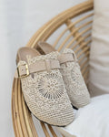 Vue de profil des mules bohèmes en crochet taupe brodé, bride à boucle carrée dorée et semelle liège, posées sur fauteuil rotin