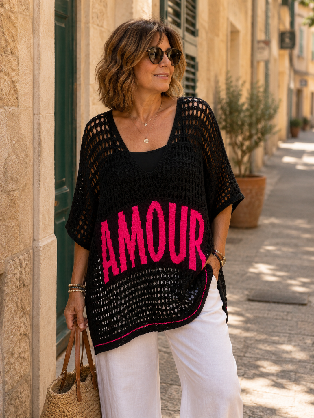 Femme 46-48 portant un pull crochet noir AMOUR style bohème avec débardeur noir, pantalon fluide blanc et lunettes de soleil dans une rue provençale lumineuse, mode italienne femme 50 ans