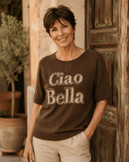 Femme portant le pull Ciao Bella chocolat en coton avec message en strass et pantalon écru devant une porte patinée en Italie