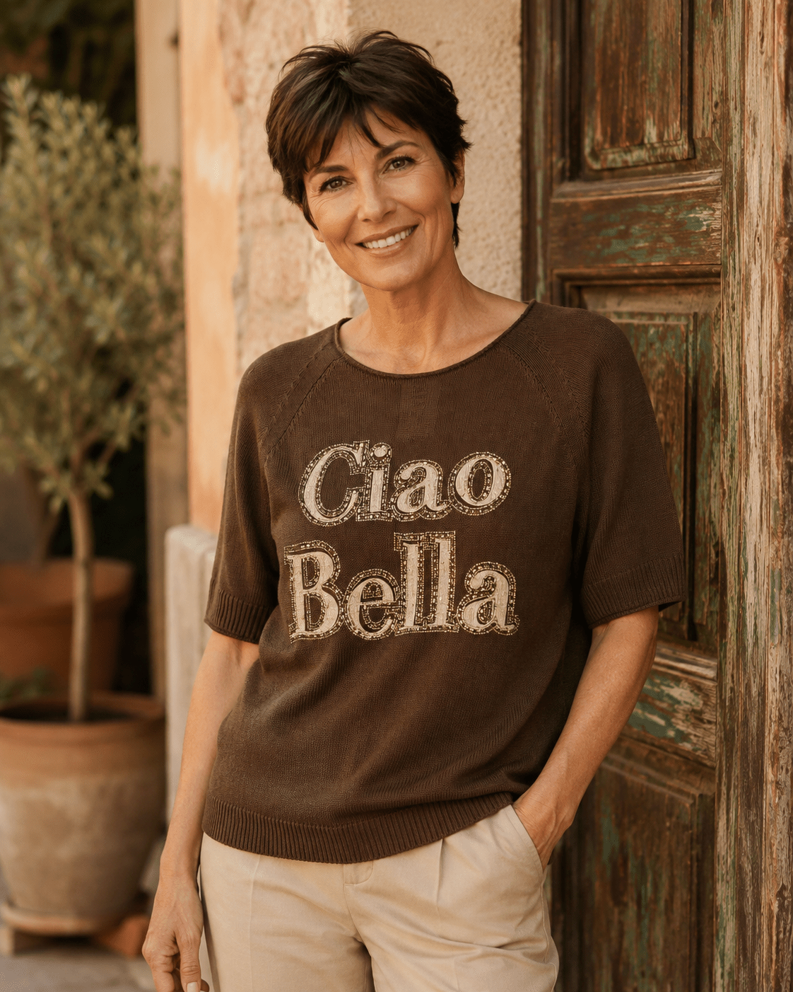 Femme portant le pull Ciao Bella chocolat en coton avec message en strass et pantalon écru devant une porte patinée en Italie
