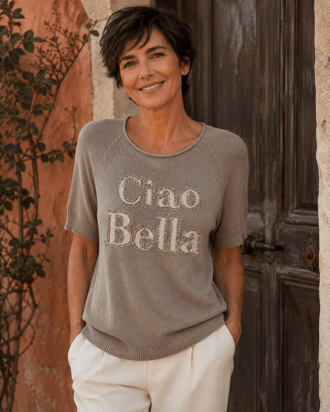 Femme portant le pull Ciao Bella taupe en coton avec message en strass et pantalon écru devant une porte ancienne en Italie