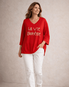 Pull rouge femme col V La Vie Bohème doré, porté avec pantalon blanc chic