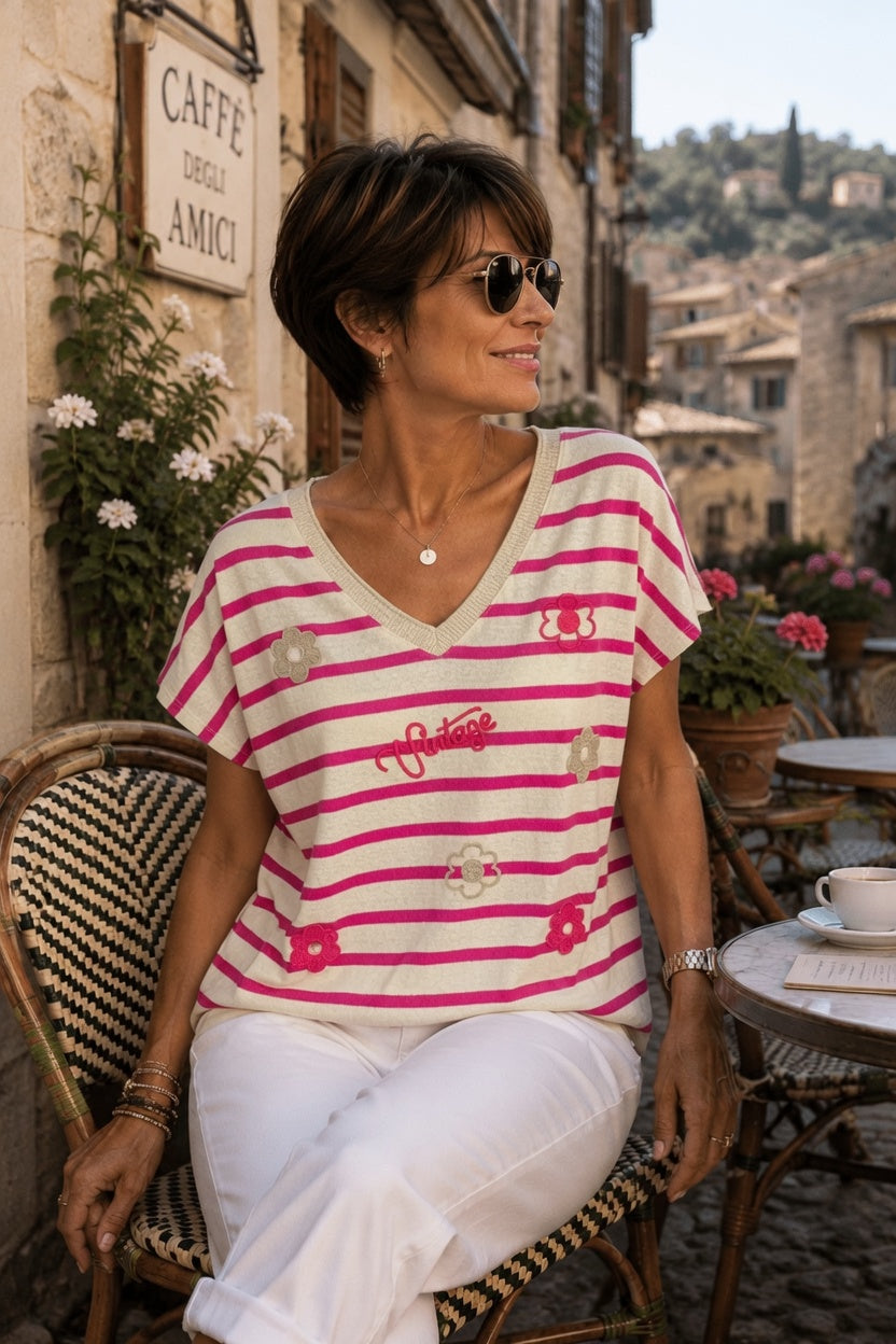 Femme 45-50 ans portant un pull rayé fushia et écru en coton italien avec motifs fleurs et inscription vintage, jean blanc sur terrasse de café italien