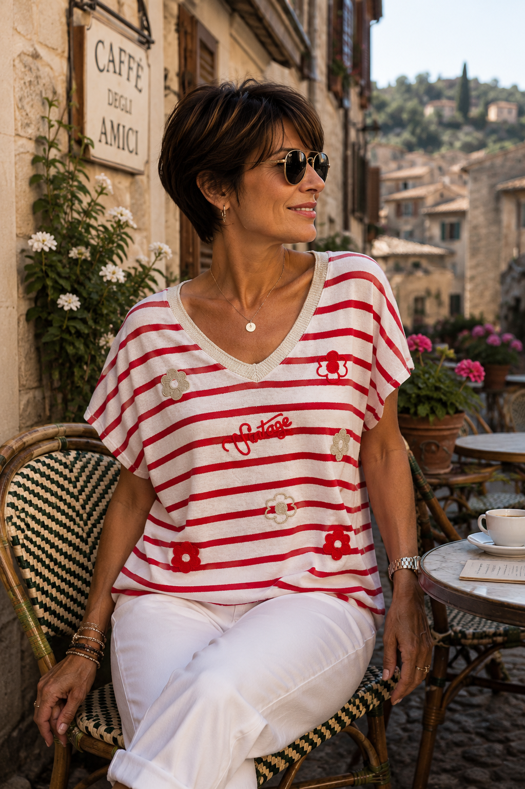 Femme brune cheveux courts portant un pull rayé rouge et écru en coton italien avec motifs fleurs vintage dans une rue de village italien près d’une terrasse café
