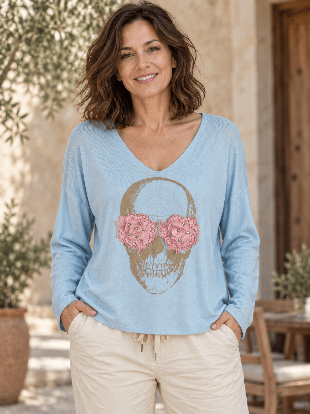 Pull bleu ciel à col V porté avec un pantalon écru, tête de mort dorée et fleurs roses en relief.
