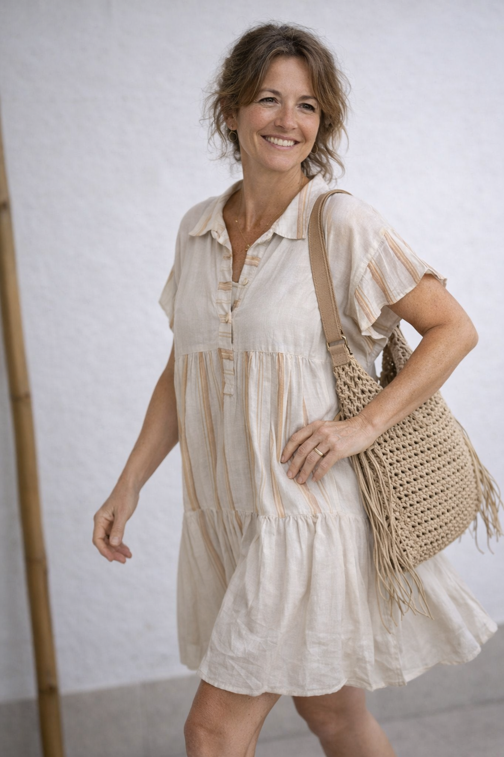 Robe bohème courte en lin écru à rayures dorées, col chemise et manches volantées, portée avec un sac crochet naturel à franges