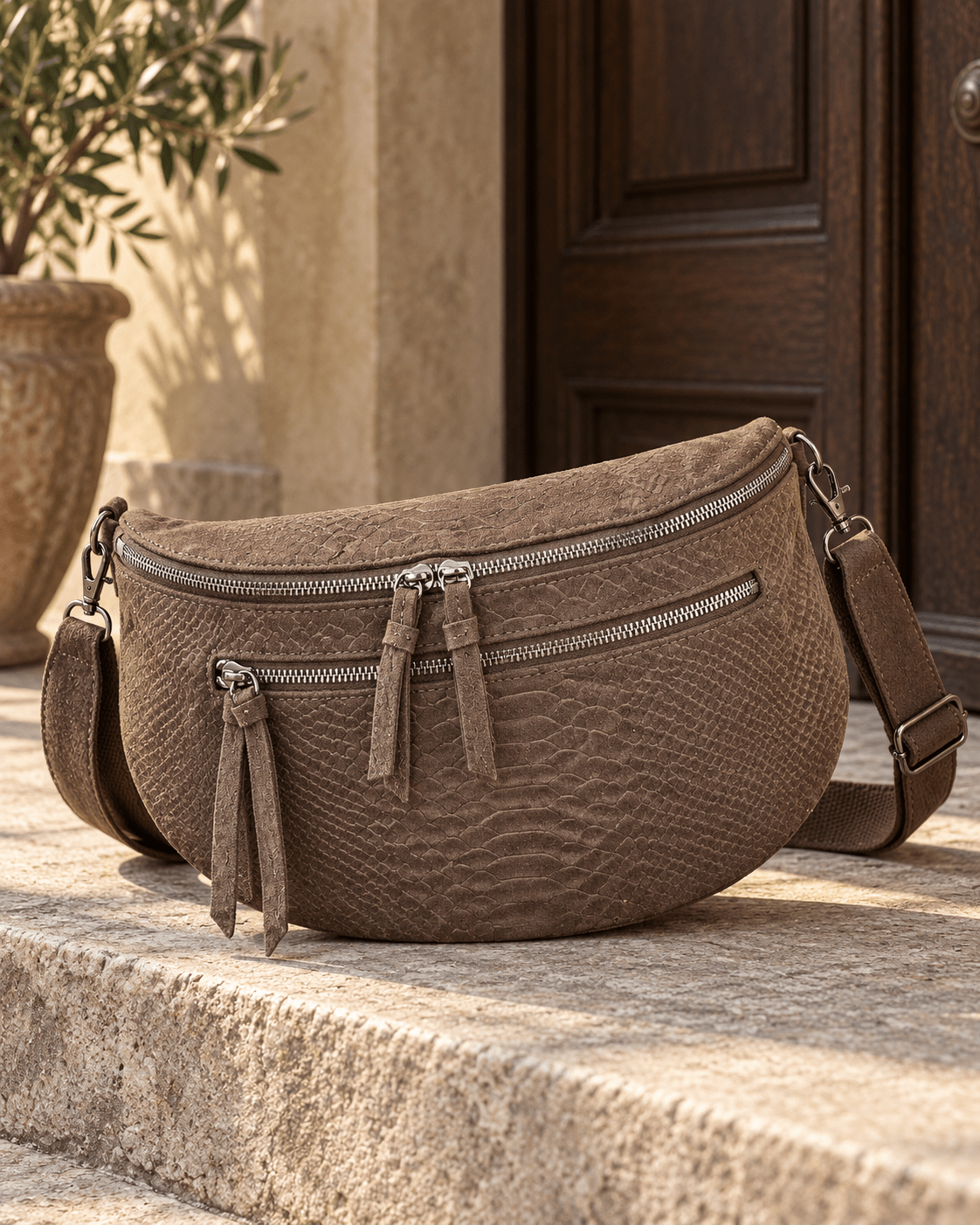 Sac banane chocolat effet serpent en simili-daim posé sur une marche en pierre, avec zips argentés et bandoulière assortie.