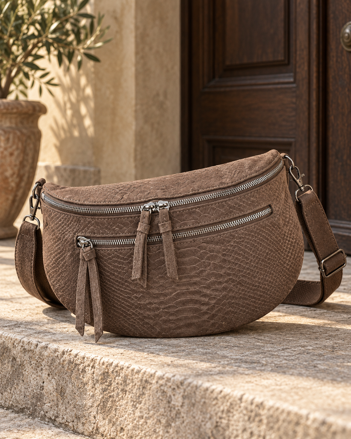 Sac banane chocolat effet serpent en simili-daim posé sur une marche en pierre, avec zips argentés et bandoulière assortie.
