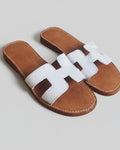 Paire de sandales slides à bride H en cuir blanc, semelle intérieure cognac surpiquée, fond gris neutre, vue 3/4 dessus