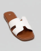 Sandale slide vue de 3/4, bride H en cuir blanc surpiqué blanc, semelle intérieure cuir cognac avec logo, fond gris épuré