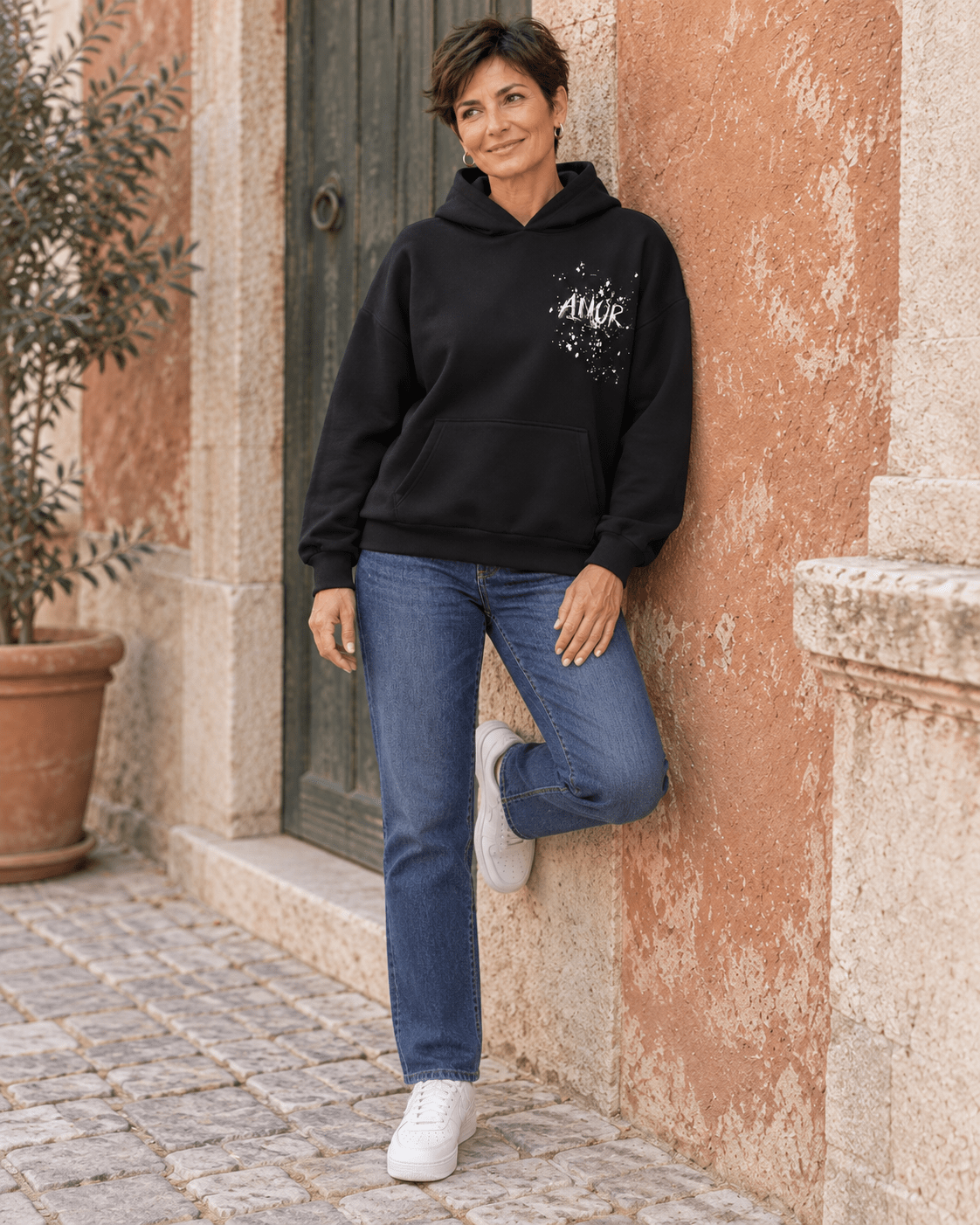 Silhouette en sweat AMOR noir et jean, adossée au mur avec une jambe pliée