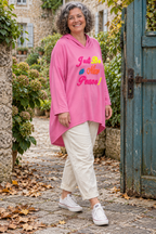 Sweat long Morgane rose Barbie porté avec un pantalon écru, capuche et inscription brodée multicolore. 