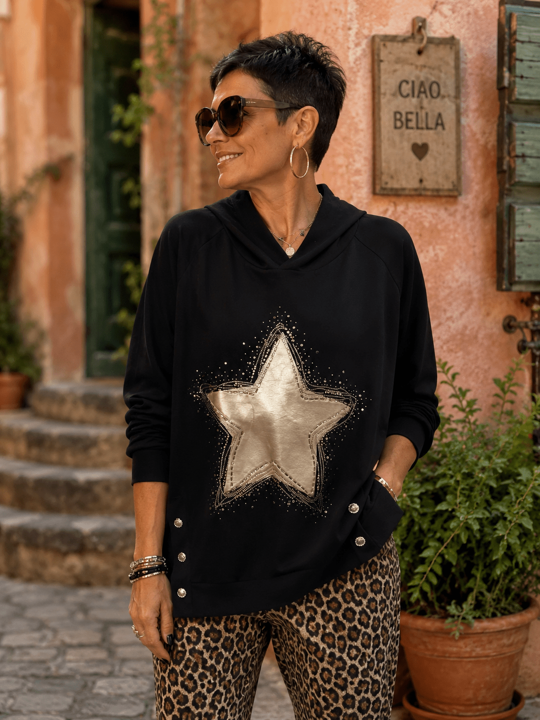 Femme portant le Sweat Luce noir à capuche avec grande étoile dorée, strass et boutons bijoux sur les côtés, dans une ruelle italienne