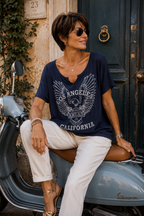 Femme 45 ans portant un tee-shirt bleu marine style rock Los Angeles California avec pantalon blanc assise sur un scooter dans une rue italienne