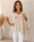 Femme de 45 à 50 ans portant une tunique bohème beige BOHEME avec pantalon blanc et sac beige à franges