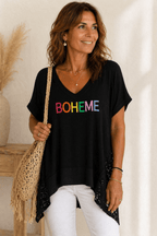 Femme de 45 à 50 ans portant une tunique noire BOHEME avec pantalon blanc, collier fin et sac macramé beige