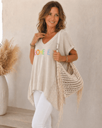 Femme de 45 à 50 ans portant une tunique beige BOHEME avec pantalon blanc, collier fin et sac macramé beige de profil