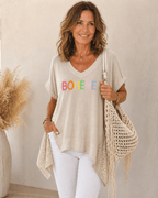 Femme de 45 à 50 ans portant une tunique noire BOHEME avec pantalon blanc, collier fin et sac macramé beige