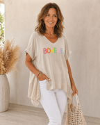 Haut beige bohème asymétrique porté par une femme bronzée avec bijoux fantaisie discrets et sac tressé