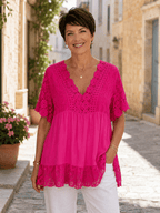 Femme portant une tunique bohème fushia en dentelle avec manches crochetées et détails floraux, style fluide chic porté avec pantalon clair dans une ruelle de village italien ensoleillée.