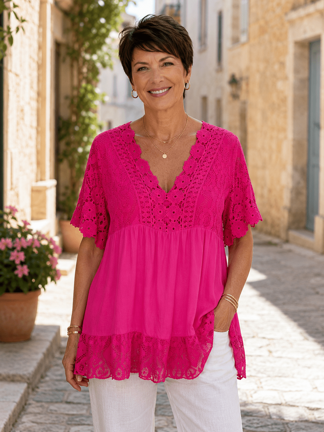 Femme portant une tunique bohème fushia en dentelle avec manches crochetées et détails floraux, style fluide chic porté avec pantalon clair dans une ruelle de village italien ensoleillée.