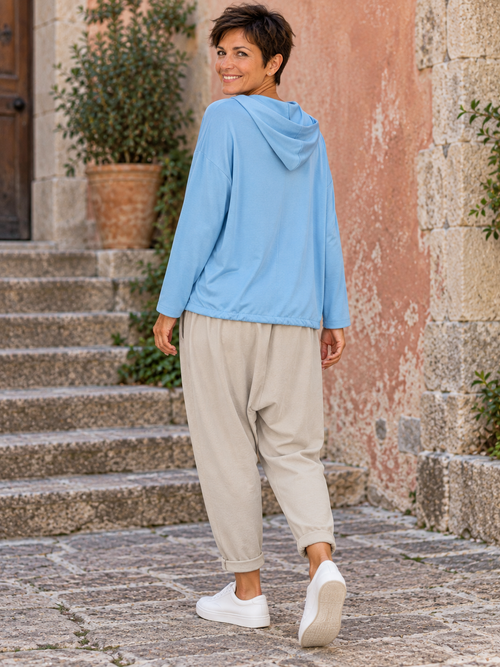 Sofia de trois quarts dos portant la veste bleu ciel à capuche, dos uni et coupe ample, avec le jogging Lou beige
